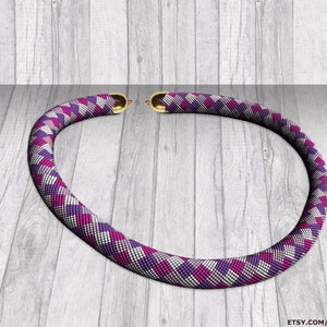 May include: A beaded necklace with a geometric pattern in shades of purple, white, and pink. The necklace has gold-colored clasps and is displayed on a light wood-grain surface. The text "BYADINDA Patterns" is visible.