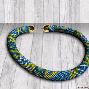 Può includere: Una collana di perline blu e gialle con chiusure dorate. La collana è realizzata con piccole perline e presenta un motivo geometrico. La collana è appoggiata su una superficie di legno.