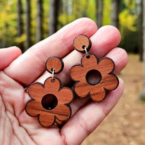 May include: A pair of wooden earrings shaped like flowers with a brown wood grain. The earrings have a simple design with a circular hole in the centre of each flower.