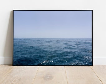 May include: A framed print featuring a seascape with a vast expanse of blue ocean under a pale blue sky. The water's surface shows gentle waves and reflections. The artwork is displayed against a white wall and light-colored wooden floor.