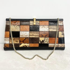 May include: A rectangular clutch bag featuring a patchwork design of various snakeskin-textured panels in shades of brown, black, tan, and cream. The bag has a solid black top and bottom border, a gold-tone metal clasp, and a gold-tone chain strap.