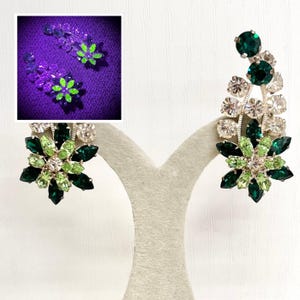 May include: A pair of silver-tone earrings featuring a floral cluster design with dark green, light green, and clear faceted rhinestones. An inset image shows the earrings under ultraviolet light, where the light green stones glow bright neon.