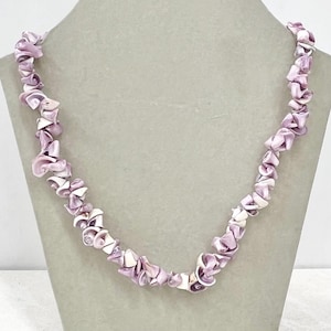 May include: A necklace made from light purple and white shell pieces, strung together to form a unique design. The shells are of irregular shapes. The necklace is displayed on a neutral-toned mannequin.