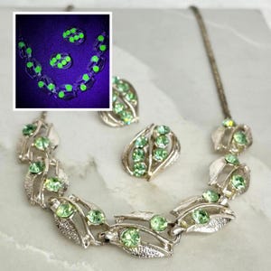 May include: A silver-toned metal jewellery set consisting of a necklace and matching clip-on earrings. Each piece features textured leaf-shaped links set with pale green round rhinestones. An inset shows the stones glowing bright neon green under UV light.