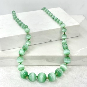 May include: A light green beaded necklace featuring large, round beads. The beads have a cat's eye effect, with a white stripe through each. The necklace is strung on a green cord and is displayed on a white surface.