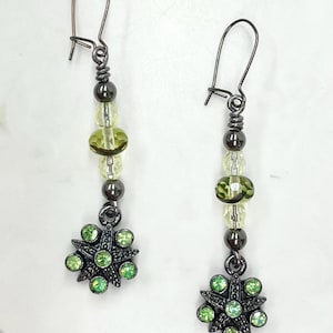 May include: A pair of drop earrings with a dark metal finish. Each earring has a hook, a series of beads in shades of green and clear, and a flower-shaped charm with green gemstones. The earrings are displayed against a white background.