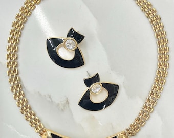 May include: A gold-toned necklace and matching earrings set. The necklace features a black enamel bar with clear, triangular accents. The earrings and necklace have a black and white design with a clear, round stone in the center. The set is on a white surface.