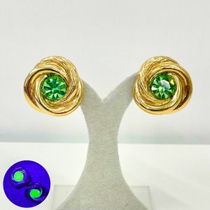 May include: A pair of gold tone earrings with green rhinestone accents. The earrings are in the shape of a knot.