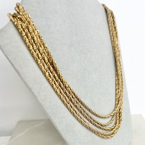 May include: A gold-coloured necklace with multiple strands, displayed on a mannequin. The necklace features a braided rope design, creating a layered effect. The warm gold colour and intricate texture are highlighted against a neutral background.