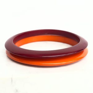 以下が含まれることがあります： 2つのバングルブレスレットが重なったセット。上のバングルは深みのある無地のバーガンディレッドで、下のバングルは鮮やかな無地のオレンジ色です。どちらのブレスレットも、滑らかで丸みを帯びた光沢のある表面をしています。