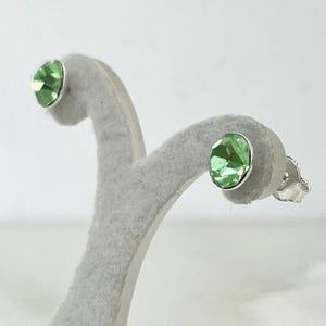 May include: Stud earrings with oval, light green gemstones set in silver-coloured metal. The earrings are displayed on a grey, felt-covered stand against a white background. The gemstones have a faceted appearance, reflecting light.