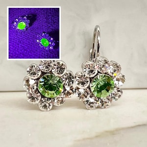 May include: A pair of leverback earrings featuring a central round light green stone surrounded by a halo of smaller clear white stones in a flower-like arrangement. An inset shows the earrings glowing bright neon green under UV light.