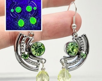 May include: A pair of silver-toned dangle earrings featuring a semi-circular metal frame set with two round green crystals and a dangling faceted light yellow bead. An inset shows the earrings glowing bright neon green under ultraviolet light.