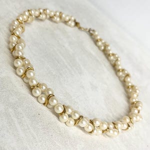 May include: A cream-colored pearl necklace with gold-tone metal accents. The necklace features multiple strands of pearls, intertwined with gold-colored metal. The clasp is also gold-toned. The necklace is displayed on a white surface.