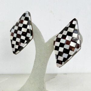 May include: A pair of diamond-shaped earrings featuring a black, white, and brown checkerboard pattern. The earrings are displayed on a light green stand against a white backdrop. The earrings have a geometric design.