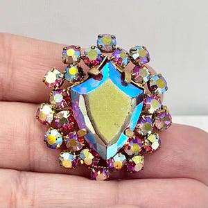 May include: A vintage brooch featuring a large iridescent faceted jewel in the shape of a shield surrounded by smaller pink and iridescent rhinestones.