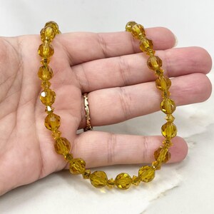 May include: A vintage necklace with amber-coloured, faceted beads. The necklace is strung with smaller, clear beads. The beads have a warm, golden hue. The necklace is held in a hand.
