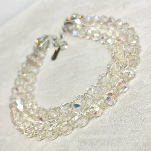 Könnte beinhalten: Ein klares, mehrsträngiges Armband mit facettierten Kristallperlen. Das Armband hat einen silberfarbenen Verschluss und wird auf einer weißen Oberfläche präsentiert. Die Kristalle reflektieren das Licht und erzeugen einen funkelnden Effekt. Dieses Schmuckstück im Vintage-Stil ist für besondere Anlässe geeignet.