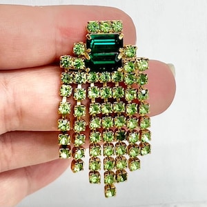 May include: A gold-tone brooch featuring a large, rectangular, dark emerald green central stone surrounded by multiple rows of smaller, light lime green square-cut rhinestones arranged in a cascading, fringe-like pattern with a metal setting.