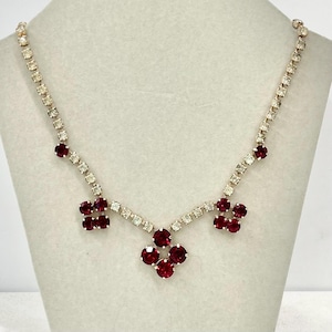 May include: A silver-toned necklace with clear crystal accents and vibrant red gemstones. The necklace features a series of clear crystals set in a delicate chain. Clusters of red gemstones are arranged in a floral pattern, creating a striking and elegant design.