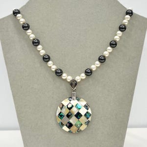 May include: A black and white pearl necklace with a round, iridescent abalone shell pendant. The pendant has a geometric pattern of squares.