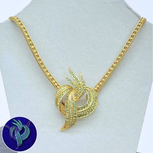 May include: A gold necklace featuring a green crystal pendant. The pendant is a knot design with a swirl pattern. The necklace is displayed against a white backdrop. The pendant glows green in the dark.