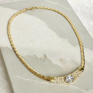 May include: A gold-toned chain necklace featuring a flat, interlocking link design. The center of the necklace showcases a curved, horizontal bar embellished with numerous small, clear crystals and a single, larger oval-shaped clear crystal.