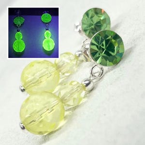 May include: Dangle earrings featuring a round, faceted lime green crystal stud connected to a silver-tone metal link and two stacked, translucent pale yellow beads. An inset shows the earrings glowing bright neon green under UV light.