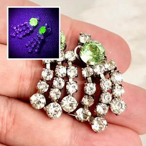 May include: A pair of chandelier earrings featuring clear round rhinestones arranged in cascading strands. Each earring is anchored by a single, larger, light green round stone. An inset shows the earrings glowing bright green under UV light.