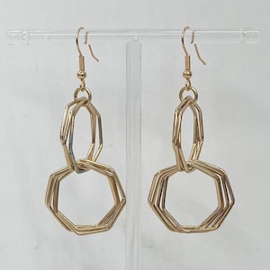 May include: A pair of gold-coloured geometric earrings. Each earring has a hook and a series of linked, multi-sided shapes. The earrings are displayed against a clear background, highlighting their design.