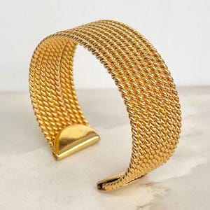 May include: A gold-toned cuff bracelet featuring a wide, textured band composed of multiple parallel rows of twisted metal wire. The design includes smooth, solid gold-toned end caps at each opening, creating a structured and uniform appearance.