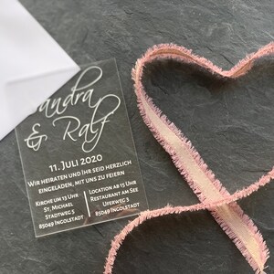 Könnte beinhalten: Eine transparente Acryl-Hochzeitseinladung mit weißer Beschriftung. Die Einladung lautet "Sandra & Ralf" und enthält das Datum "11. Juli 2020". Die Einladung enthält auch die Details für die Zeremonie und den Empfang, einschließlich des Ortes, der Uhrzeit und der Adresse.