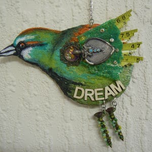 May include: A green, blue and orange bird-shaped ornament with the word "DREAM" written on it. The ornament is decorated with a metal charm, beads and a piece of yellow measuring tape.