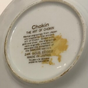K&ouml;nnte beinhalten: Eine wei&szlig;e dekorative Platte mit goldenem Rand, die Text &uuml;ber Chokin-Kunst aus Japan zeigt. Der Text beschreibt mentale Gravuren, die mit Gold und Silber vergoldet wurden, urspr&uuml;nglich f&uuml;r die Bewaffnung der Samurai-Krieger. Die Platte ist mit "24K GOLD EDGED" und "MADE IN JAPAN" gekennzeichnet.