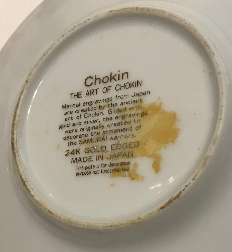 K&ouml;nnte beinhalten: Eine wei&szlig;e dekorative Platte mit goldenem Rand, die Text &uuml;ber Chokin-Kunst aus Japan zeigt. Der Text beschreibt mentale Gravuren, die mit Gold und Silber vergoldet wurden, urspr&uuml;nglich f&uuml;r die Bewaffnung der Samurai-Krieger. Die Platte ist mit "24K GOLD EDGED" und "MADE IN JAPAN" gekennzeichnet.