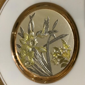 K&ouml;nnte beinhalten: Ein wei&szlig;er Teller mit Goldrand und einem zentralen Medaillon. Das Medaillon zeigt ein detailliertes Relief eines Kolibris und Blumen in Silber- und Goldt&ouml;nen, wodurch ein dekoratives Design entsteht.