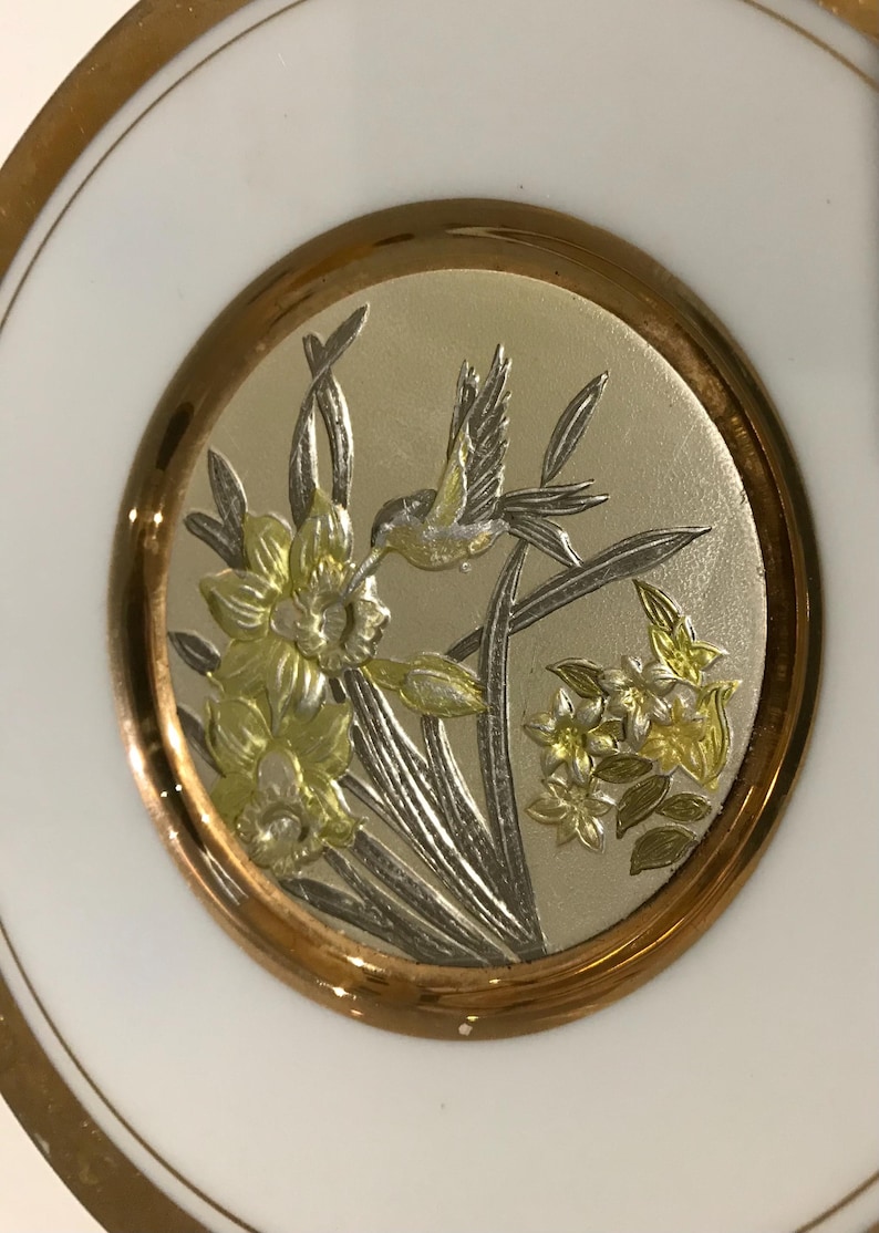 K&ouml;nnte beinhalten: Ein wei&szlig;er Teller mit Goldrand und einem zentralen Medaillon. Das Medaillon zeigt ein detailliertes Relief eines Kolibris und Blumen in Silber- und Goldt&ouml;nen, wodurch ein dekoratives Design entsteht.