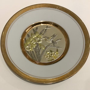 K&ouml;nnte beinhalten: Ein runder, wei&szlig;er Teller mit goldenem Rand. In der Mitte befindet sich ein goldenes und silbernes Design eines Kolibris und Blumen. Der innere Kreis hat einen goldenen Rand. Der Teller ist ein dekorativer Gegenstand.