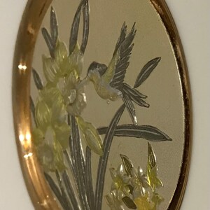K&ouml;nnte beinhalten: Ovales Dekorationselement mit goldfarbenem Rand. Die Mitte zeigt eine detaillierte Illustration eines Kolibris in der N&auml;he von gelben und wei&szlig;en Blumen. Der Hintergrund ist hell und neutral, und das Gesamtdesign ist elegant.