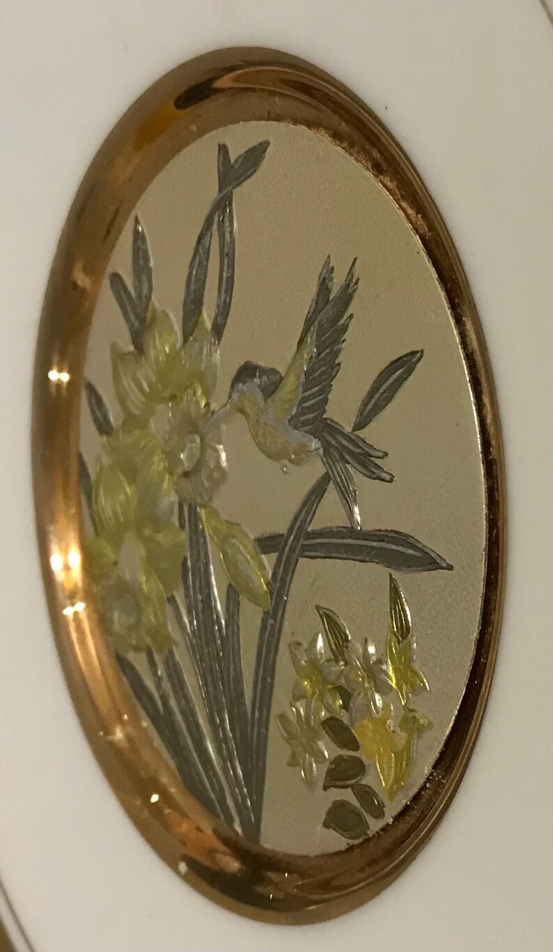 K&ouml;nnte beinhalten: Ovales Dekorationselement mit goldfarbenem Rand. Die Mitte zeigt eine detaillierte Illustration eines Kolibris in der N&auml;he von gelben und wei&szlig;en Blumen. Der Hintergrund ist hell und neutral, und das Gesamtdesign ist elegant.
