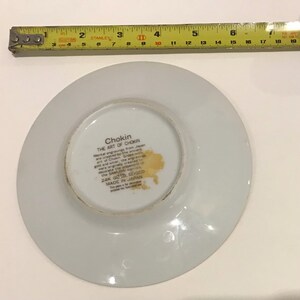 K&ouml;nnte beinhalten: Eine wei&szlig;e Chokin-Platte mit Goldrand, mit dem Text "Chokin THE ART OF CHOKIN" und "24K GOLD EDGED MADE IN JAPAN". Ein gelbes Ma&szlig;band liegt &uuml;ber der Platte und zeigt Messungen in Zentimetern.