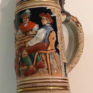 May include: A ceramic beer stein with a pewter lid and handle. The stein depicts a scene of two men seated at a table, enjoying a drink. The stein is decorated with floral patterns and has a gold trim.