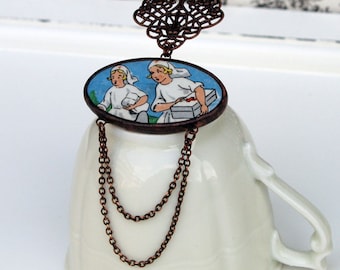 Pode incluir: Um colar com pingente de estilo vintage com uma imagem oval de duas figuras em roupas brancas sobre um fundo azul. O pingente é suspenso por uma corrente de cor bronze com um elemento decorativo de filigrana. O colar é exibido em uma xícara de chá branca.