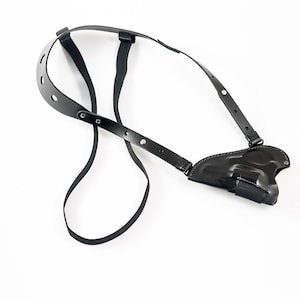 Könnte beinhalten: Ein schwarzes Leder-Schulterholster für eine Pistole. Das Holster verfügt über verstellbare Riemen und einen Druckknopfverschluss.