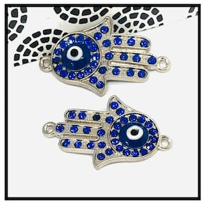 Può includere: Due ciondoli a forma di mano di Hamsa color argento con accenti di strass blu e un design centrale a occhio blu. I ciondoli hanno piccoli anelli per il fissaggio a gioielli o altri lavori artigianali. Sfondo bianco.