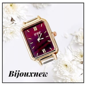 Peut inclure: Une montre dorée avec un cadran carré et un cadran bordeaux. La montre a des index en chiffres romains et un bracelet doré.
