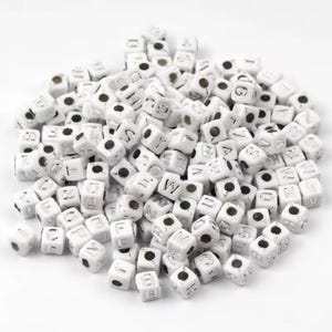 Peut inclure: Un grand tas de perles en plastique blanches avec des lettres argentées. Les perles sont en forme de cube et ont un trou au centre.