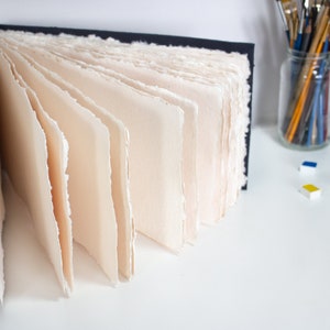 Puede incluir: Un cuaderno de bocetos azul marino con p&aacute;ginas en blanco de color crema. Las p&aacute;ginas est&aacute;n hechas a mano y tienen un borde rugoso y texturizado. El cuaderno de bocetos est&aacute; abierto y desplegado, revelando las p&aacute;ginas del interior.