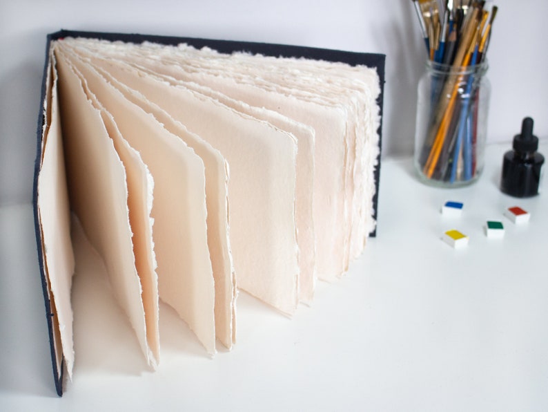 Puede incluir: Un cuaderno de bocetos azul marino con p&aacute;ginas en blanco de color crema. Las p&aacute;ginas est&aacute;n hechas a mano y tienen un borde rugoso y texturizado. El cuaderno de bocetos est&aacute; abierto y desplegado, revelando las p&aacute;ginas del interior.