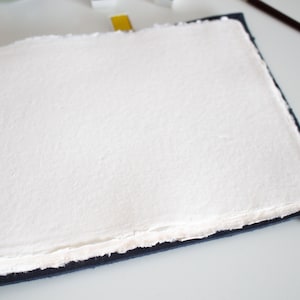 Puede incluir: Una hoja de papel blanco texturizado, en blanco, encuadernada en una cubierta azul oscuro. El papel tiene un borde irregular.